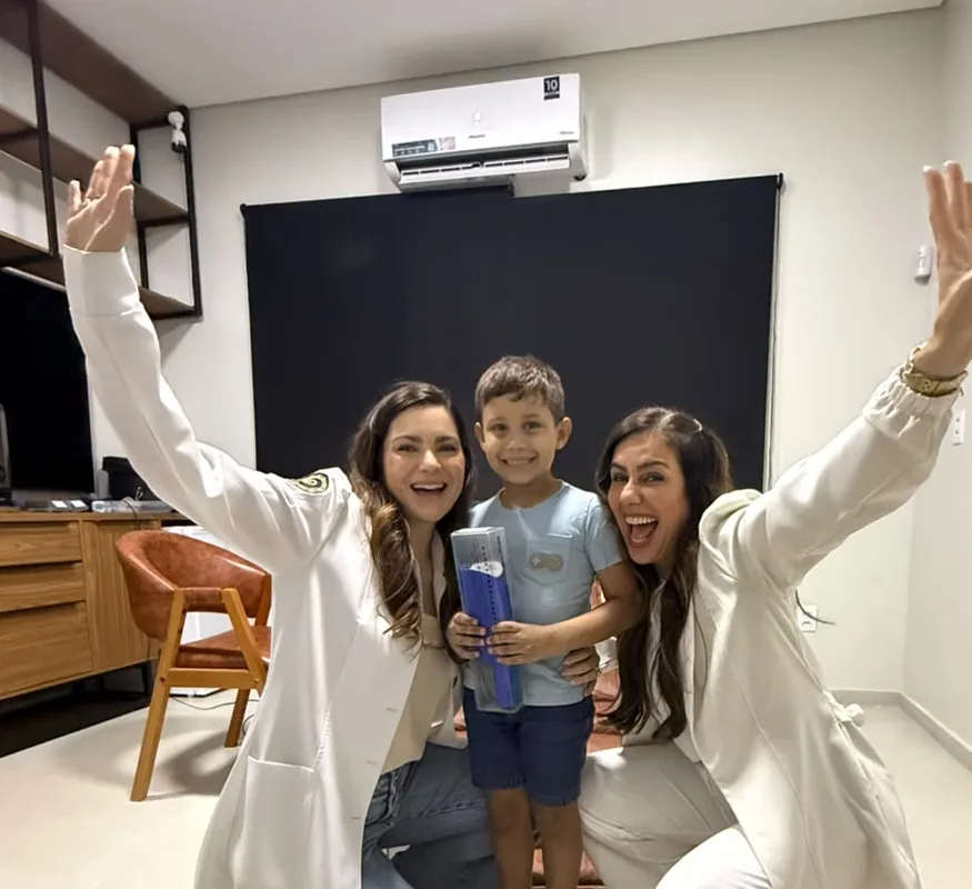 Menino segurando o seu kit entre as duas profissionais, que comemoram de braços para cima